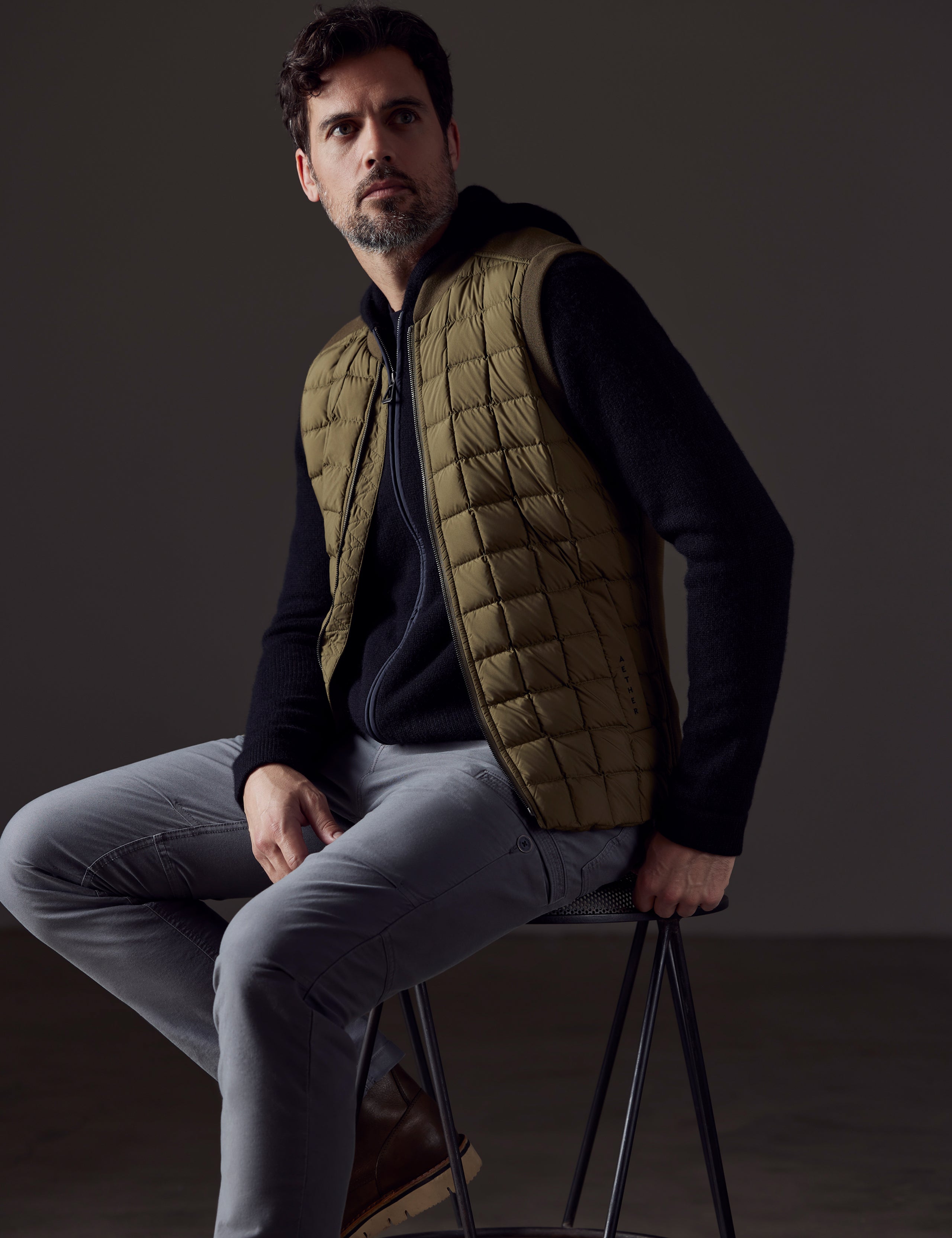 Front 3/4 body view of man sitting wearing Phase Vest in Command Green from AETHER Apparel.