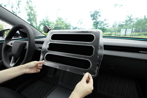A person holds a JOWUA Tempered Glass Screen Protector for the Tesla Model 3 Highland 2024+ in a sleek vehicle, with the dashboard and steering wheel visible and trees and buildings framed through the windshield—perfect for this model.