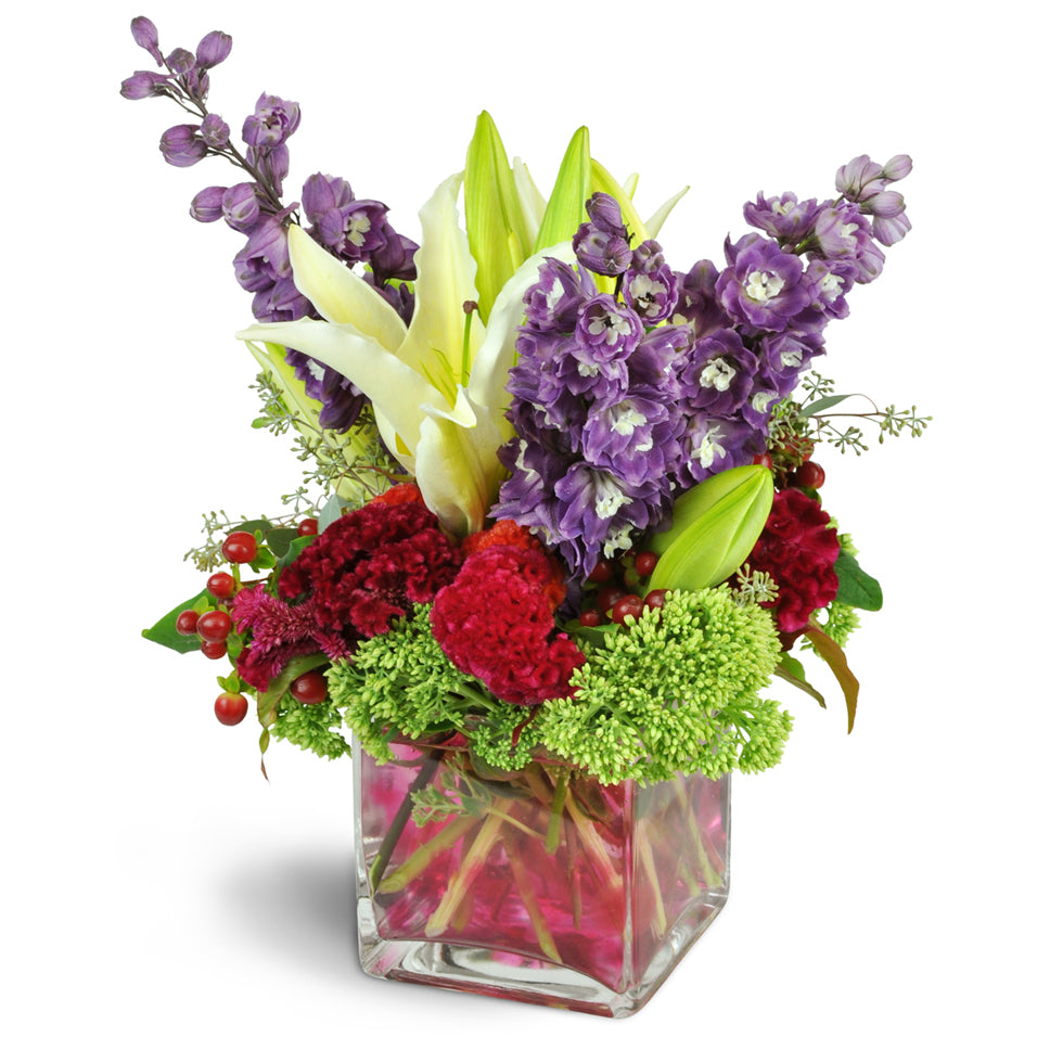 Magnificent Jewel™. Lilies, delphinium, bells of Ireland, and more, all in luxe jewel tones, are arranged in a modern clear glass vase.
