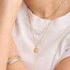 Person wearing a white shirt with the Pave Diamond Bail Round Tag Charm Pendant, featuring a 14k gold round tag and pavé diamond-set bail.