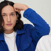 A man with long hair wearing a blue jacket and a Figaro chain necklace, posing with one hand on his forehead.